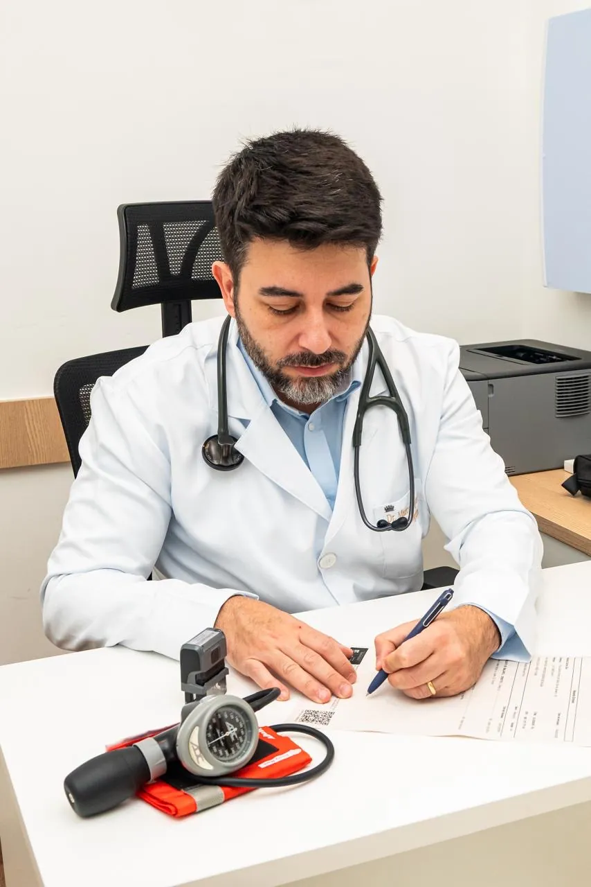Dr. Marcus Vinicius Iglesias de estetoscópio, redigindo uma prescrição no consultório